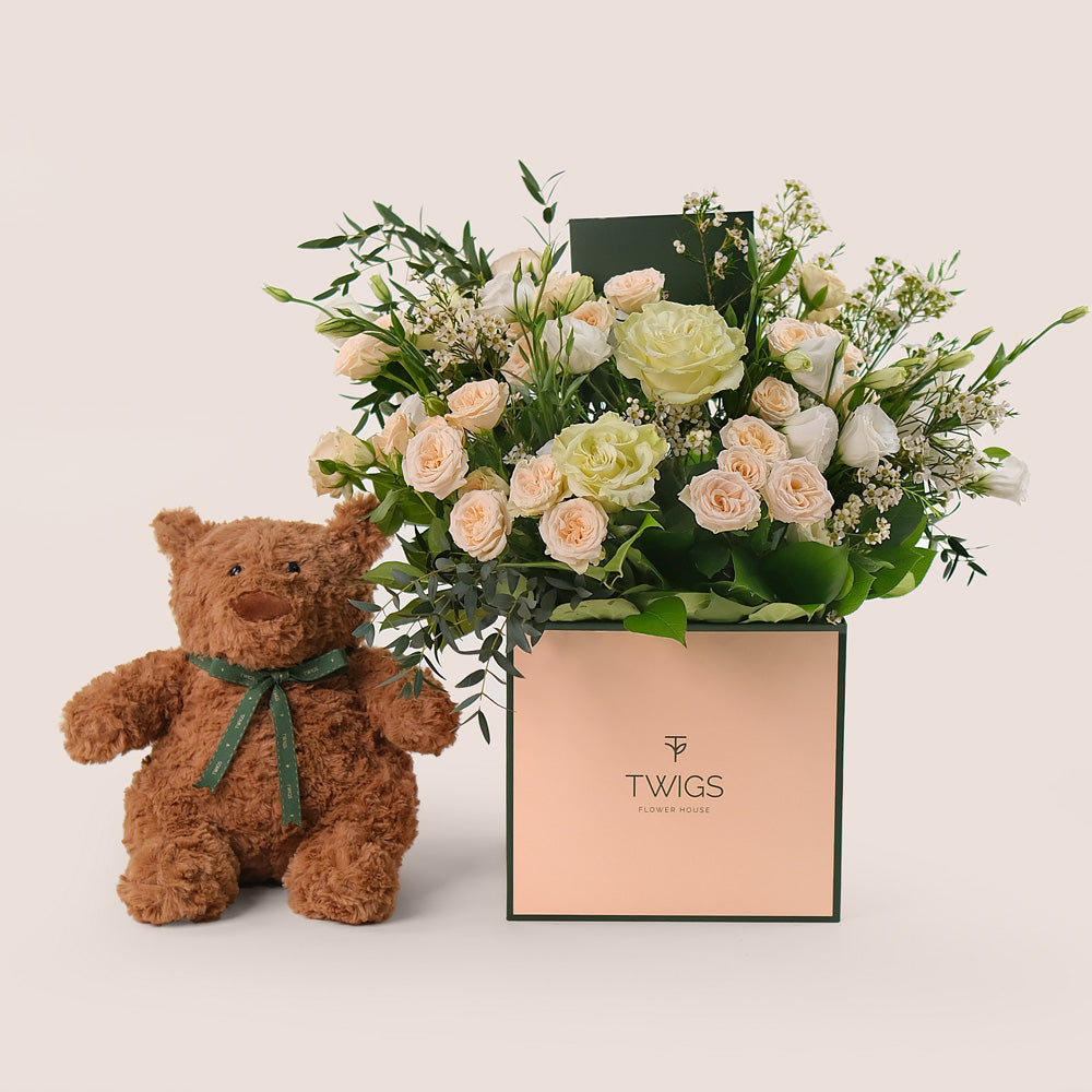 A beige square flower box with a arrangement of white and cream flowers, accompanied by a plush teddy bear with a green ribbon around its neck.