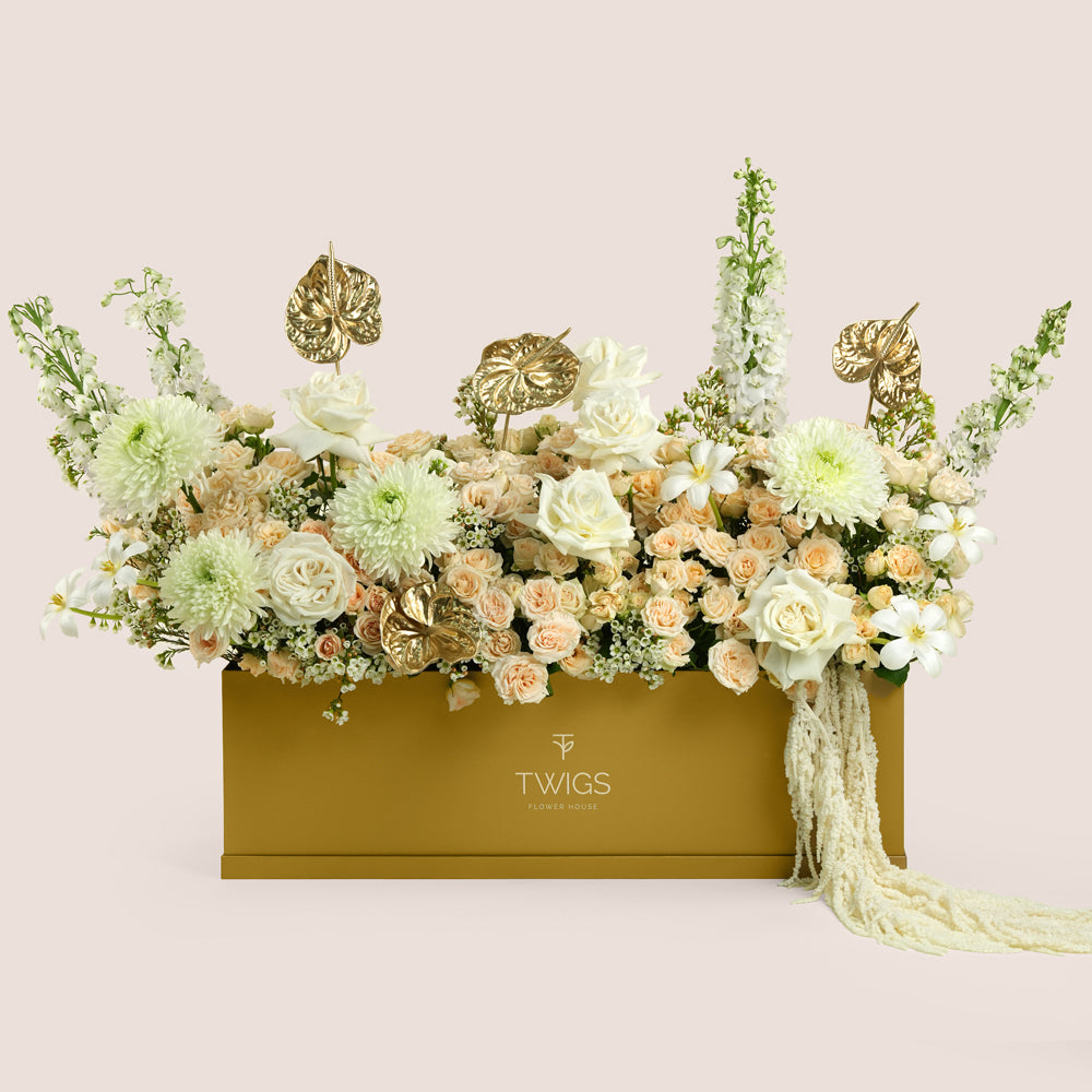 Lumina, a luxury floral arrangement of white roses, peach spray roses, chrysanthemums, delphiniums, and gold painted anthurium leaves in a rectangular TWIGS Gold garden box, with a long cream amaranthus accent.