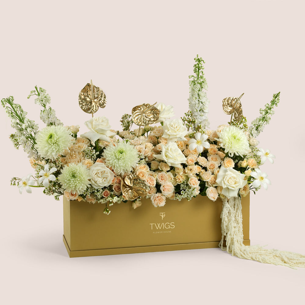 Lumina, a luxury floral arrangement of premium roses, peach spray roses, chrysanthemums, delphiniums, and gold painted anthurium leaves in a rectangular TWIGS Gold garden box, with a long cream amaranthus accent.