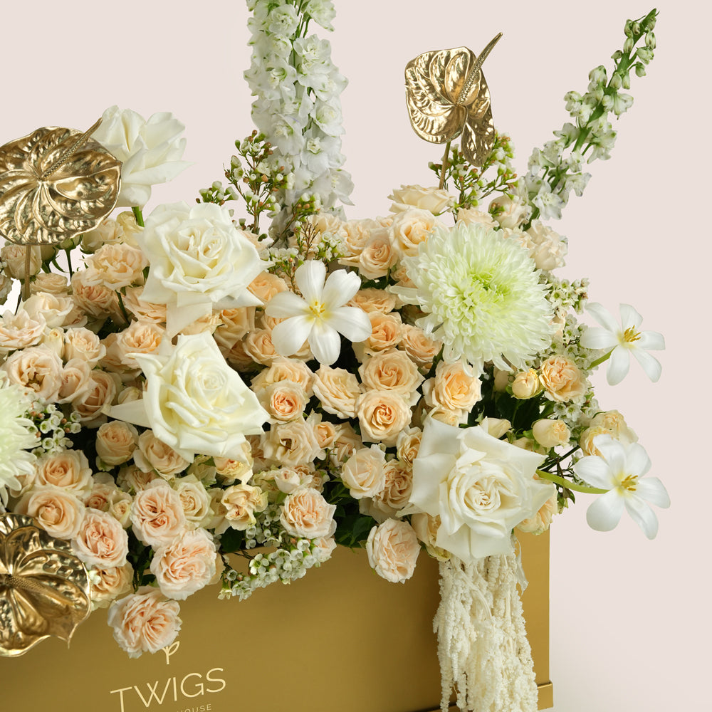 Lumina, a luxury floral arrangement of white roses, peach spray roses, chrysanthemums, delphiniums, and gold painted anthurium leaves in a rectangular TWIGS Gold garden box, with a long cream amaranthus accent.