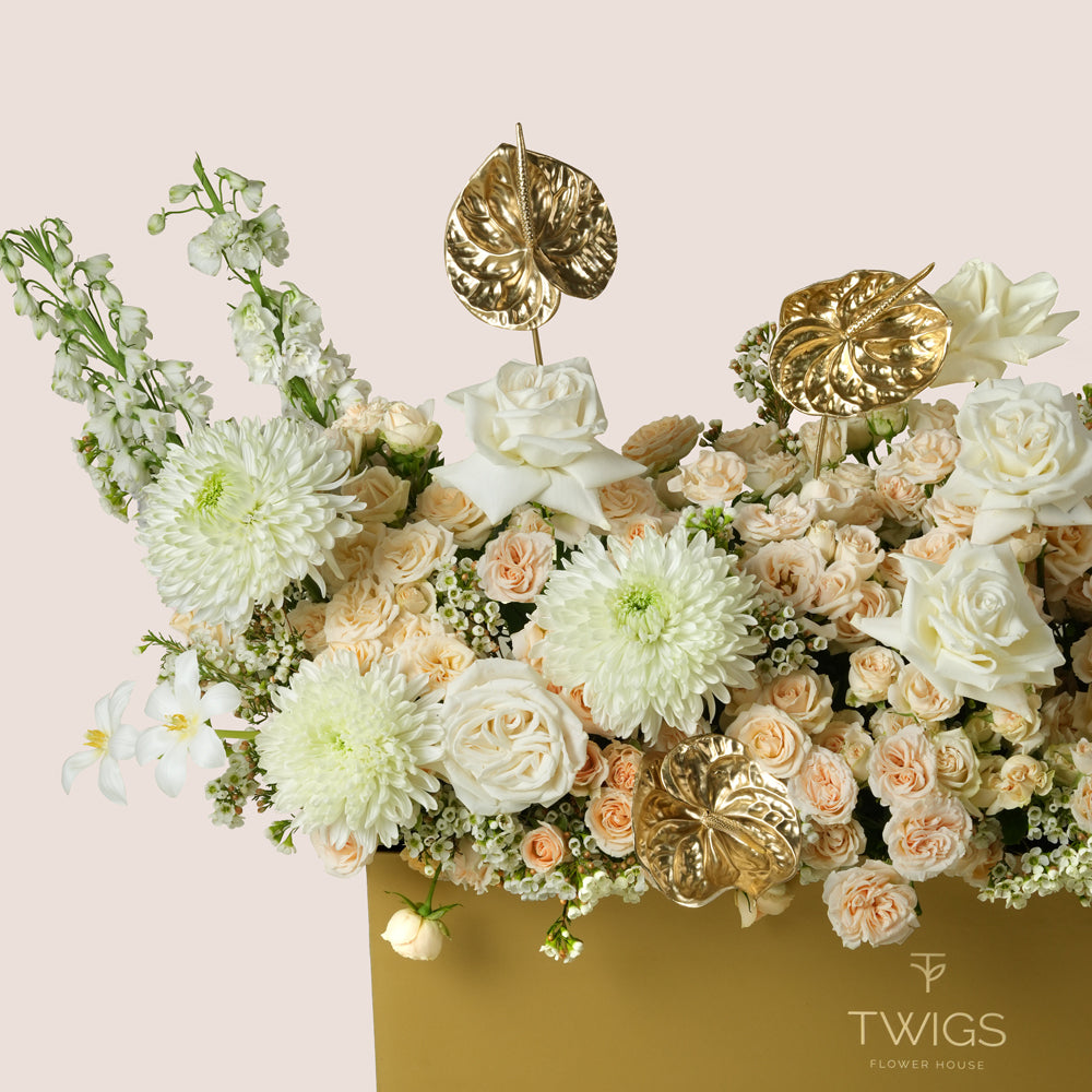 Lumina, a luxury floral arrangement of white roses, peach spray roses, chrysanthemums, delphiniums, and gold painted anthurium leaves in a rectangular TWIGS Gold garden box, with a long cream amaranthus accent.