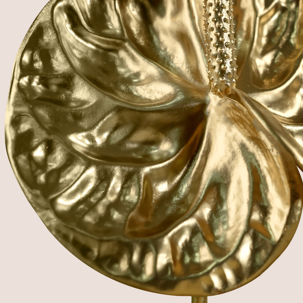 A close-up of a metallic gold anthurium leaf ornament with a textured hanging loop at the top, set against a solid light beige background.