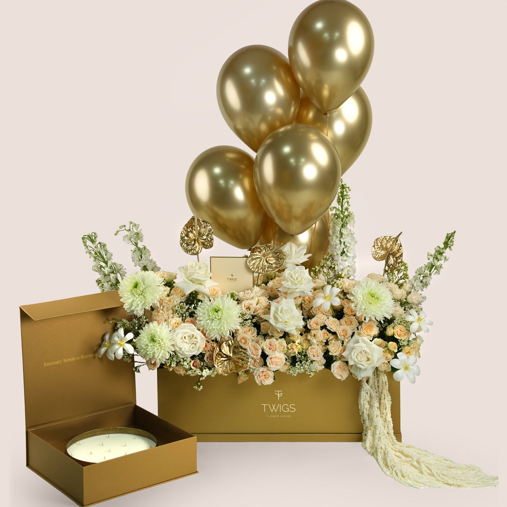Lumina, a luxury TWIGS Gold Garden box arrangement of garden roses, chrysanthemums, and gold anthurium leaves, paired with the Palace Candle by Light of Sakina and six gold chrome balloons.