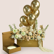 Lumina, a luxury TWIGS Gold Garden box arrangement of garden roses, chrysanthemums, and gold anthurium leaves, paired with the Palace Candle by Light of Sakina and six gold chrome balloons.