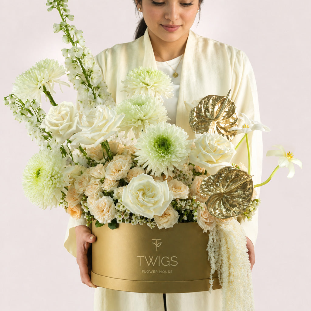 Lumina, a luxury floral arrangement of white garden roses, deco chrysanthemums, salinero spray roses, delphiniums, tulips, and gold painted anthurium leaves in a round TWIGS Gold box, with matching gift card.