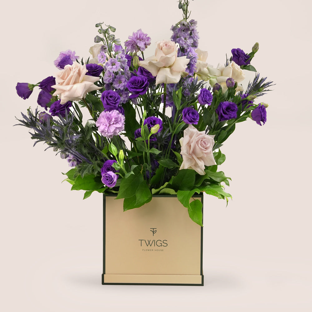 Square flower box with quicksand roses, purple lisianthus, light purple carnations, delphiniums, and eryngium in TWIGS beige or green packaging, available for same day flower delivery in Dubai when you buy flowers online.
