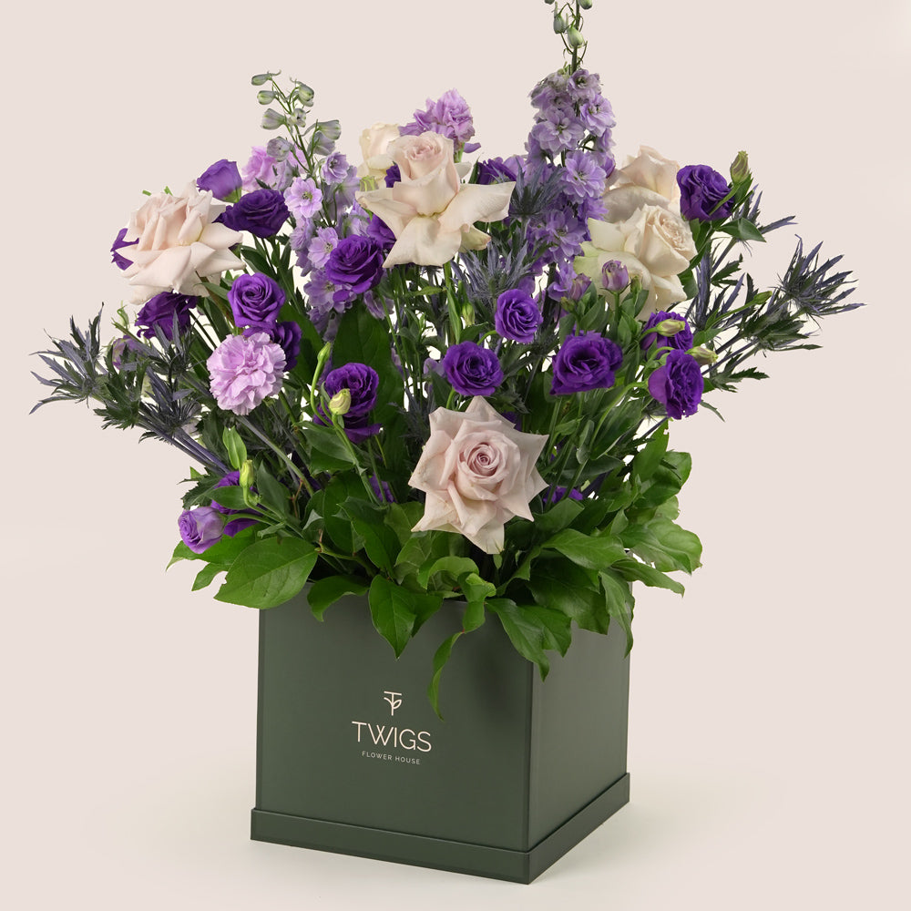 Square flower box with quicksand roses, purple lisianthus, light purple carnations, delphiniums, and eryngium in TWIGS beige or green packaging, available for same day flower delivery in Dubai when you buy flowers online.