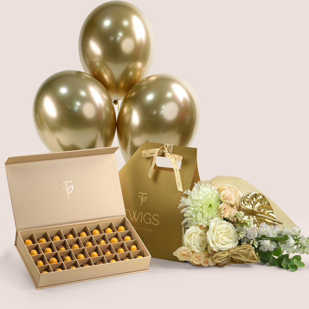 Lumina Indulgence, a TWIGS Gold carry bag with white roses, chrysanthemums, and a gold anthurium leaf accent, paired with a 32 piece premium bonbon box and three gold chrome balloons.